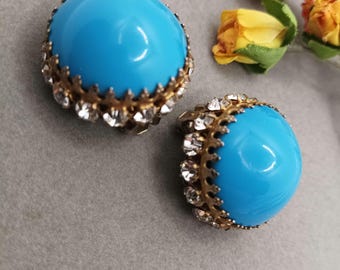 Puede incluir: Un par de pendientes vintage de clip. Cada pendiente presenta una piedra cabujón redonda y brillante de color azul claro, engastada en un marco de metal dorado. El marco está adornado con pequeños cristales transparentes. Los pendientes se muestran sobre una superficie gris.