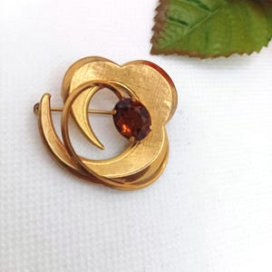 Puede incluir: Broche dorado con diseño floral. El broche presenta una gema central, de forma ovalada, de color marrón rojizo. El diseño incluye elementos curvos en capas y un cierre de alfiler en la parte posterior. El broche está sobre un fondo blanco.