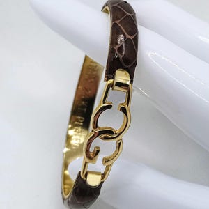 May include: A brown leather bracelet with a gold metal clasp featuring interlocking 'C's. The bracelet has the text 'PIERRE CARDIN' engraved on the metal band.