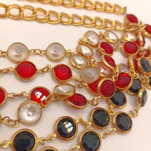 May include: Close-up of several gold-toned necklaces with various gemstone accents. The necklaces feature red, clear, and black circular stones set in gold bezels. A thicker gold chain is visible at the top. The jewelry is displayed against a white background.