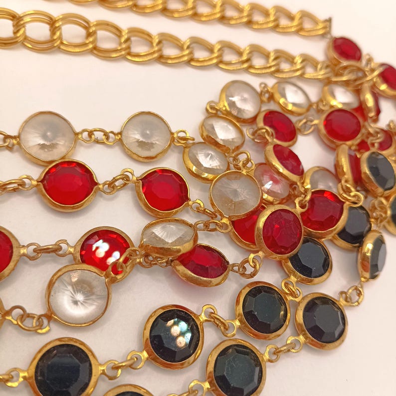 May include: Close-up of several gold-toned necklaces with various gemstone accents. The necklaces feature red, clear, and black circular stones set in gold bezels. A thicker gold chain is visible at the top. The jewelry is displayed against a white background.