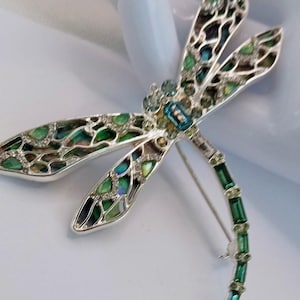 Op de afbeelding: Zilveren libellebroche met groen en blauw iriserende accenten op de vleugels. De broche is versierd met heldere kristallen en groene rechthoekige stenen. Een stijlvol accessoire.