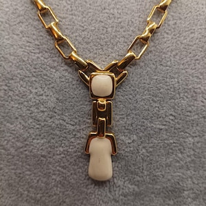 May include: Gold-tone necklace with a geometric design. The necklace features a chain with rectangular links and a pendant with white, rectangular and rounded elements. The pendant has a vertical drop design.