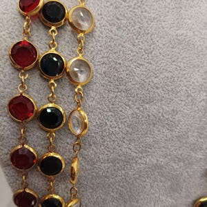 May include: Close-up of a gold-toned necklace with three strands of large, round gemstones. The gemstones are red, black, and clear, set in gold bezels. The necklace is displayed against a gray background.
