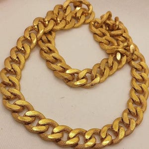 Op de afbeelding: Een goudkleurige ketting met dikke, in elkaar grijpende schakels met een getextureerde, matte afwerking. De ketting is in een cirkelvorm gelegd, waardoor de uniforme grootte en het herhalende patroon van de metalen schakels zichtbaar zijn.