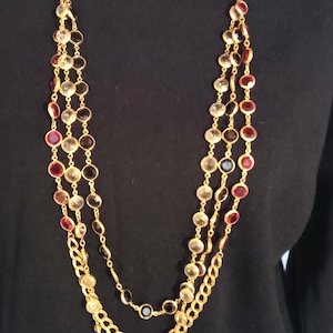May include: A multi-strand necklace with gold-tone chains and three strands of alternating clear, red, and black circular stones. The necklace is displayed against a black background.
