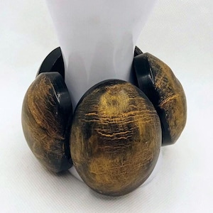 Può includere: Un bracciale elastico massiccio composto da grandi maglie ovali e arrotondate. Ogni maglia presenta una base di colore nero scuro con una finitura dorata testurizzata e usurata sulla superficie anteriore, creando un aspetto ad alto contrasto.