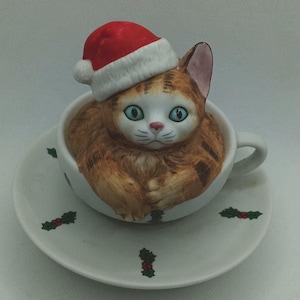 May include: A ceramic figurine of a cat wearing a Santa hat, nestled inside a teacup. The cat has orange and white fur and blue eyes. The teacup and saucer are white with holly berry accents.