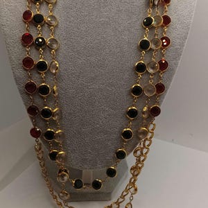 May include: A multi-strand necklace with gold chains and alternating colored gemstones. The gemstones are red, black, and clear, set in gold bezels. The necklace is displayed on a mannequin.
