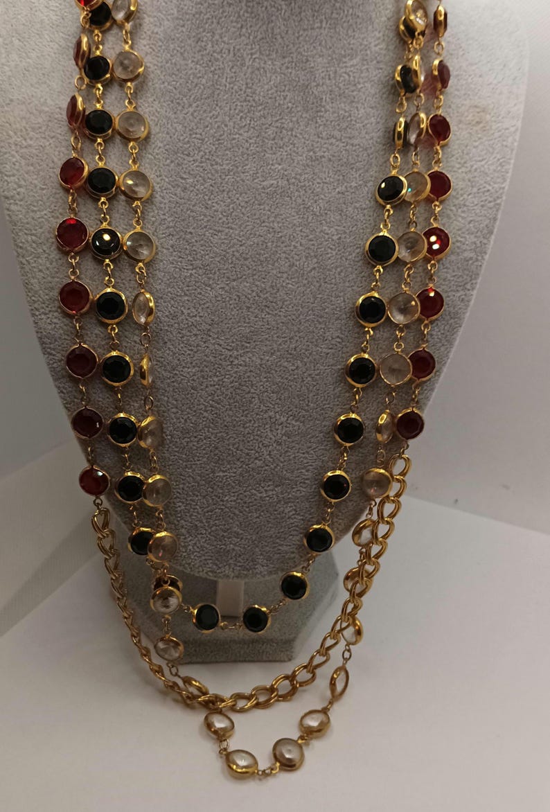 May include: A multi-strand necklace with gold chains and alternating colored gemstones. The gemstones are red, black, and clear, set in gold bezels. The necklace is displayed on a mannequin.