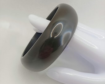 Może przedstawiać: Ciemnobrązowa, błyszcząca bransoletka typu bangle. Gładka, zaokrąglona bransoletka jest prezentowana na białym, ręcznie ukształtowanym wyświetlaczu. Bransoletka jest w jednolitym kolorze i ma prosty, elegancki design. Bransoletka to klasyczny dodatek.