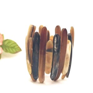 May include: A cuff bracelet constructed from multiple vertical, elongated, smooth pieces of material in varying shades of brown, tan, cream, and dark charcoal gray, arranged in a dense, overlapping pattern to form a cylindrical shape.