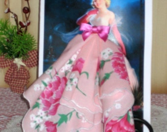 Peut inclure: Un mouchoir en dentelle rose à fleurs avec un nœud en satin rose noué autour. Le mouchoir est plié pour ressembler à une robe et est présenté devant une carte avec une image d'une femme en robe rose.