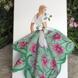 May include: A white card with a drawing of a woman wearing a white dress with a green and pink floral handkerchief attached to the back of the dress.