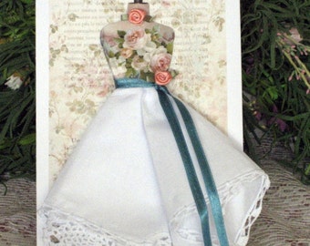Peut inclure: Carte de vœux faite à la main avec un mannequin miniature orné de motifs floraux. Le mannequin est orné de roses roses et d'un ruban turquoise. La jupe est faite d'un mouchoir blanc avec une bordure en dentelle.