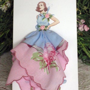 May include: A vintage-style greeting card featuring a fashion illustration of a woman in a blue dress with pink floral accents. The dress is crafted from a pink and blue handkerchief, adding a unique, handmade touch. The card is displayed on a small gold stand.