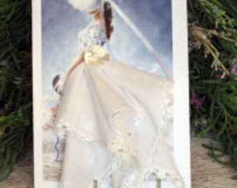 Peut inclure: Carte de vœux faite à la main représentant une mariée en robe blanche et voile. La mariée porte un chapeau et tient un bouquet. Le fond est un motif aquarelle bleu et blanc. La carte est encadrée en blanc.