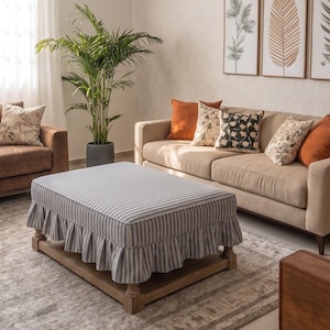 Op de afbeelding: Een rechthoekige poef met een grijs en wit gestreepte stoffen hoes met een geplooide rok, rustend op een houten frame met vier korte poten. De poef staat voor een beige bank die is versierd met oranje en bedrukte kussens.