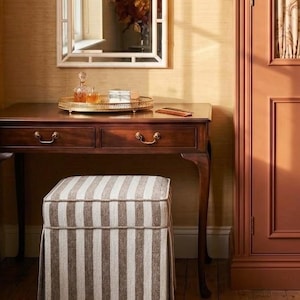 Op de afbeelding: Een donkerhouten sidetable met twee lades en messing handgrepen, gecombineerd met een vierkante, gestoffeerde poef met verticale beige en witte strepen. Een rond rieten dienblad bevat een glazen karaf en een klein doosje onder een spiegel.