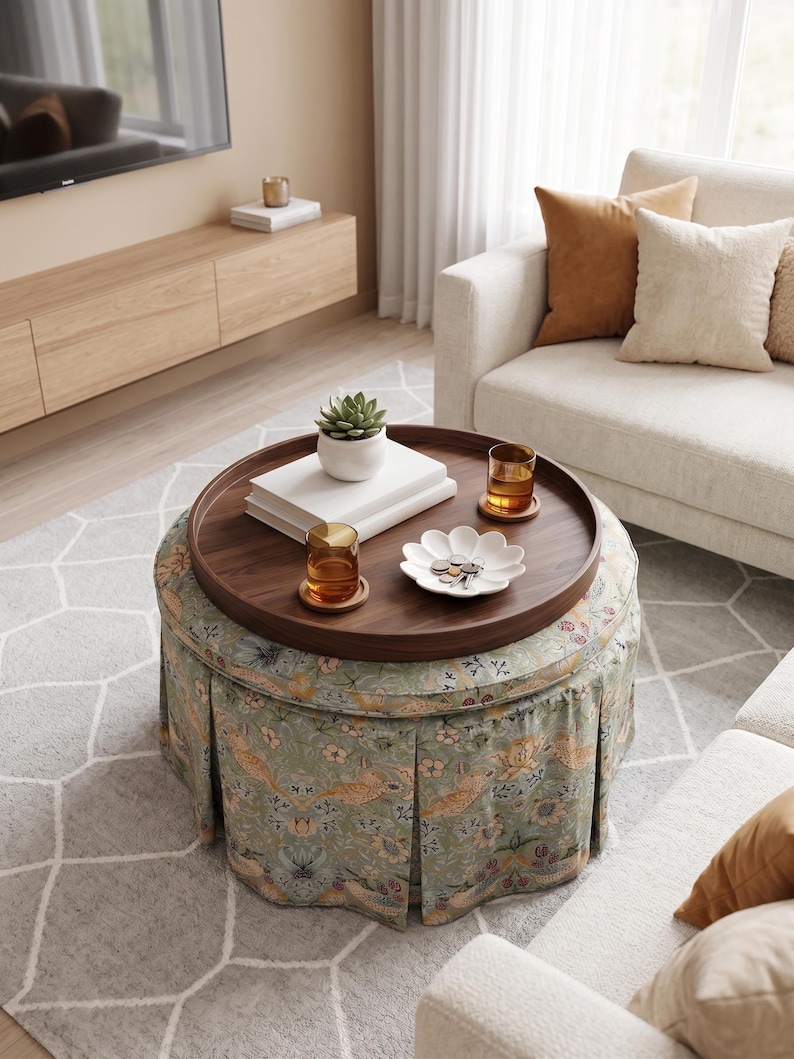 May include: A round upholstered ottoman featuring a pleated skirt with a light green floral and bird pattern. A circular dark wood tray sits on top, holding two amber glasses, a white succulent planter, stacked books, and a white flower-shaped dish.