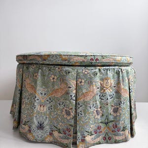 May include: A round ottoman featuring a pleated skirt upholstered in a light blue fabric with a repeating pattern of birds, flowers, and berries in shades of tan, pink, and dark blue. The top cushion is circular with matching fabric piping.