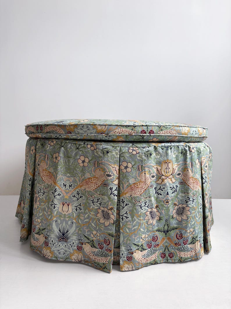 May include: A round ottoman featuring a pleated skirt upholstered in a light blue fabric with a repeating pattern of birds, flowers, and berries in shades of tan, pink, and dark blue. The top cushion is circular with matching fabric piping.