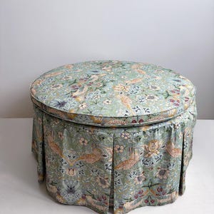 May include: A round ottoman featuring a light green fabric with a repeating pattern of birds, flowers, and foliage. The piece has a cushioned top and a pleated skirt that extends to the base, covering the structure in a consistent floral print.