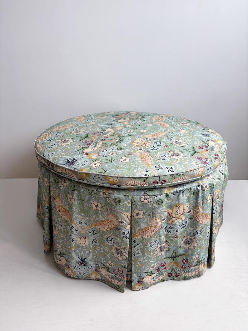 May include: A round ottoman featuring a light green fabric with a repeating pattern of birds, flowers, and foliage. The piece has a cushioned top and a pleated skirt that extends to the base, covering the structure in a consistent floral print.