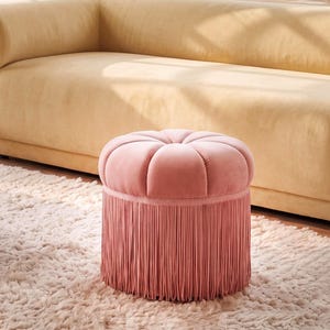 May include: A round, dusty pink velvet ottoman featuring a tufted top with a central button and a base adorned with long, matching pink fringe trim that extends to the floor, creating a soft, textured appearance for this circular footstool.