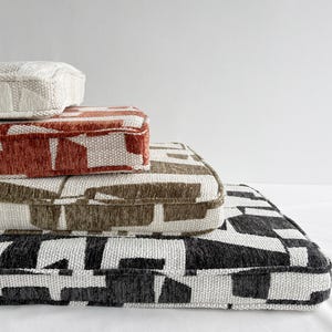 May include: Four stacked rectangular cushions with geometric patterns. The top cushion is off-white, followed by rust, brown, and black and white. The cushions have a textured appearance and are arranged on a white surface.