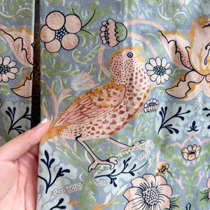 May include: A light blue fabric featuring a repeating pattern of stylized birds in shades of peach and brown, surrounded by intricate floral motifs in soft pink, green, and dark blue. The textile has a visible woven texture and a matte finish.