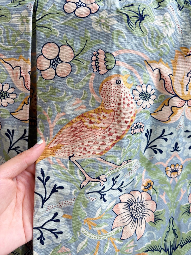 May include: A light blue fabric featuring a repeating pattern of stylized birds in shades of peach and brown, surrounded by intricate floral motifs in soft pink, green, and dark blue. The textile has a visible woven texture and a matte finish.