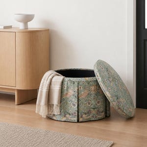 May include: A round, upholstered storage ottoman featuring a green fabric with a detailed floral and bird pattern. The lid is detached and resting beside the base. A beige, fringed throw blanket is draped over the side of the cylindrical container.