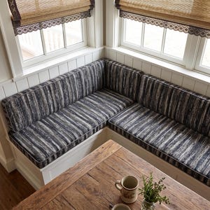 May include: An L-shaped corner bench featuring white wooden base cabinetry and thick, upholstered cushions with a dark gray, black, and white vertical striped pattern. The bench is positioned against a white wall with windows above.