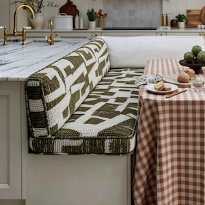 May include: A built-in kitchen bench with a green and white geometric patterned cushion. A white bolster pillow rests against the back. A brown and white checkered tablecloth covers a table set with food and a decorative bowl.