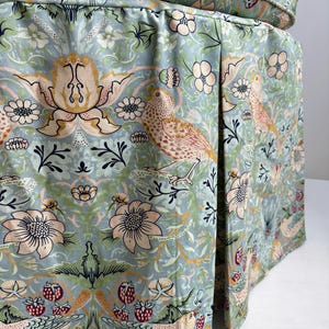 May include: A round ottoman featuring a light blue fabric cover with a repeating pattern of birds, flowers, and strawberries in shades of pink, yellow, and green. The base includes a pleated skirt design with a matching circular top cushion.