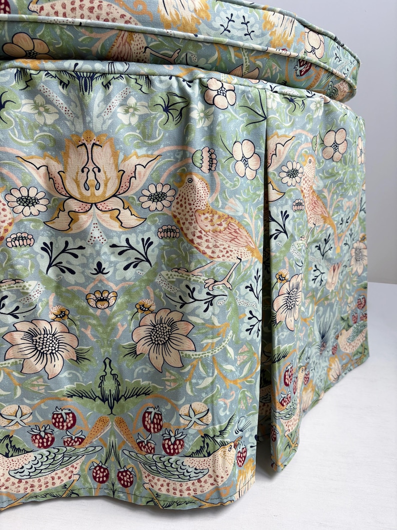 May include: A round ottoman featuring a light blue fabric cover with a repeating pattern of birds, flowers, and strawberries in shades of pink, yellow, and green. The base includes a pleated skirt design with a matching circular top cushion.