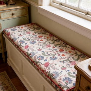 May include: A cream-coloured storage bench with a floral cushion. The cushion has a cream background with blue, pink, and orange flowers and green leaves. A red trim borders the cushion. The bench is near a window.