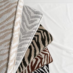 May include: A stack of decorative pillows with zebra stripe patterns in various colors. The pillows include beige, gray, black, brown, and olive green with white accents. The pillows are arranged on a white surface.