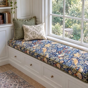May include: A white built-in window seat with four drawers and brass knobs, topped with a long cushion featuring a blue, yellow, and green floral and bird pattern. Two rectangular throw pillows, one solid olive green and one cream with botanical prints, rest on the cushion.