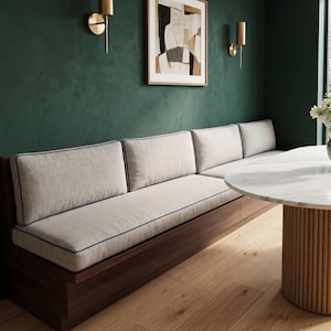 May include: A long wooden bench seat with a dark brown base and light beige upholstered cushions. The cushions feature thin blue piping along the edges. The bench is positioned against a dark green wall and paired with a round white marble table.