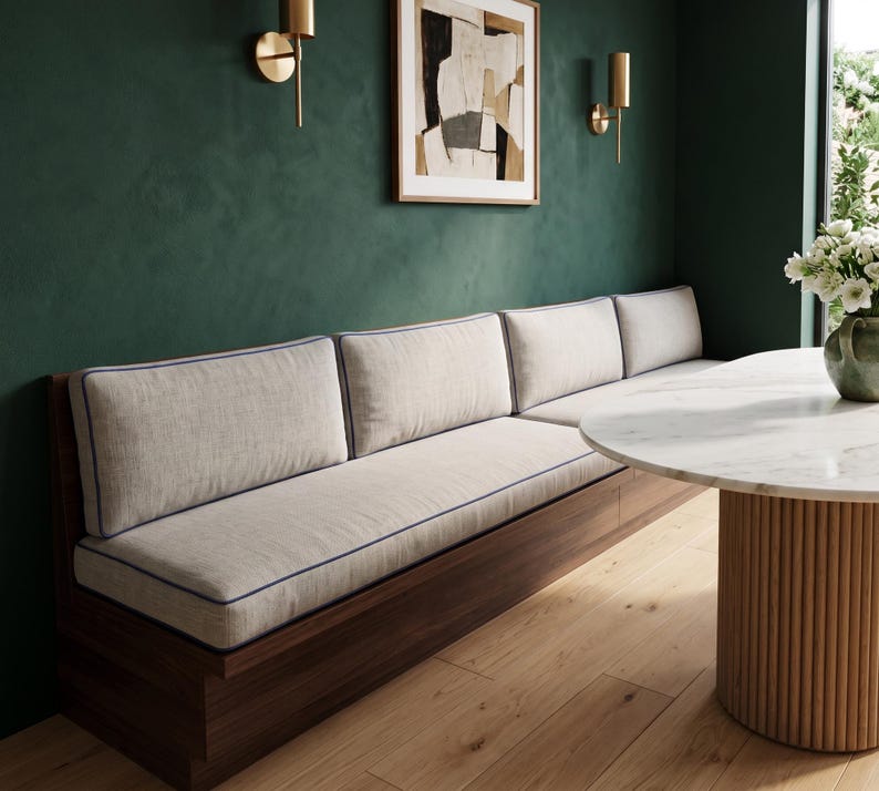 May include: A long wooden bench seat with a dark brown base and light beige upholstered cushions. The cushions feature thin blue piping along the edges. The bench is positioned against a dark green wall and paired with a round white marble table.