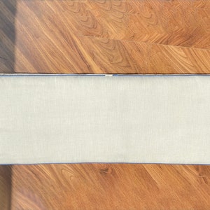 May include: A rectangular bench cushion featuring a light beige, textured fabric cover. The cushion is finished with a vibrant blue piping trim that outlines the entire perimeter, providing a clean and defined edge to the rectangular shape.