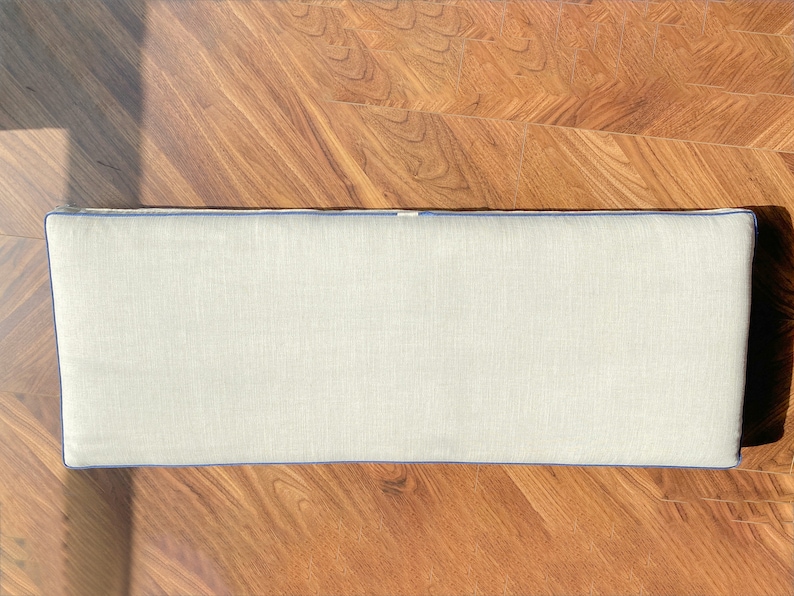May include: A rectangular bench cushion featuring a light beige, textured fabric cover. The cushion is finished with a vibrant blue piping trim that outlines the entire perimeter, providing a clean and defined edge to the rectangular shape.