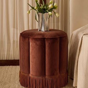 May include: A round, rust-coloured velvet ottoman featuring vertical channel tufting and a thick, matching fringe trim along the base. A silver-toned, fluted vase containing a bouquet of white tulips sits on the flat top surface of the ottoman.