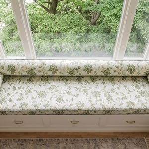 May include: A long rectangular window seat cushion with two matching cylindrical bolster pillows, all upholstered in a cream fabric featuring a repeating green floral pattern. The cushion and bolsters are positioned on a white built-in bench.