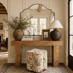 May include: A rectangular wooden console table with thick legs, topped with a large dark ceramic vase containing green branches, a stack of books, two metal candle holders, a framed picture, and a dark ceramic lamp with a white shade.