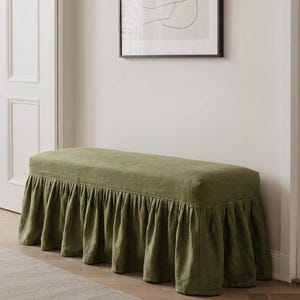 May include: A rectangular bench upholstered in olive green fabric featuring a long, gathered skirt that extends to the floor. The bench has a flat, cushioned top surface and a continuous pleated fabric valance covering the base on all visible sides.