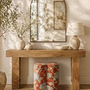 May include: A rectangular wooden console table with thick legs, topped with two ceramic vases, a bowl, and a table lamp. An arched mirror hangs above the table. Below the table sits a four-lobed stool upholstered in a rust-colored fabric with a floral pattern.