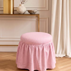 May include: A round, light pink ottoman featuring a textured fabric upholstery and a gathered, floor-length skirt. The seat is circular with a piped edge, and the base is fully covered by the pleated fabric drape, creating a soft, uniform appearance.